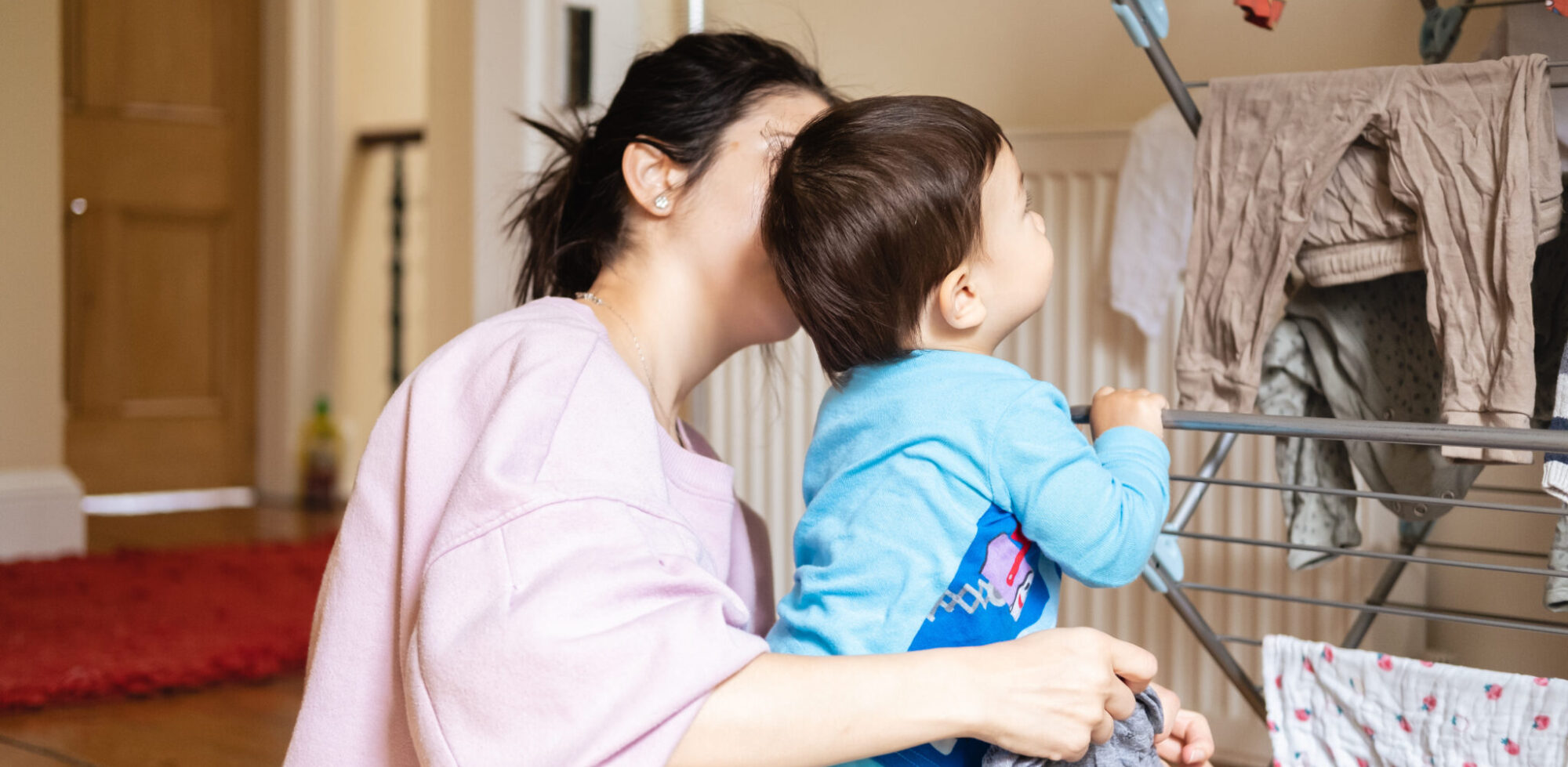 A Unrecognizable New Mother Trying To Hang Out The Laundry While Her Son, A Male Infant, Gets In The Way. Real Mixed Race Family At Home In Pajamas.